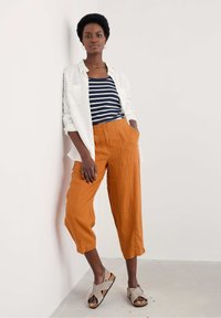 Orange linnebyxor i cropped längd med fickor, kombinerade med en marinblå och vit randig linne topp och en vit skjorta. Grå sandaler fulländar looken.