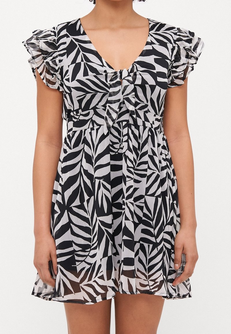 Femme portant une robe courte noire et blanche à motif feuilles, avec des manches courtes volantées et un lien à nouer sur le devant.