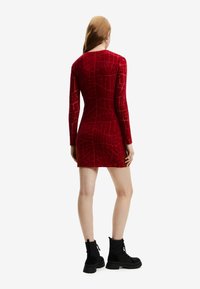 Rotes Samt-Kleid mit langen Ärmeln und einem strukturierten geometrischen Muster, taillierte Silhouette und Oberschenkel-Länge, kombiniert mit schwarzen Knöchelschuhen.