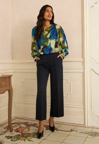 Hosszú, sötét hajú nő kék, zöld és fekete mintás blúzt visel, amelyet fekete, bő szárú, térd alatti nadrágba tűr, benti környezetben áll.