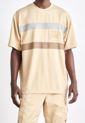Beige short-sleeve t-shirt featuring horizontal gray and brown stripes. Material appears textured. Logo with a lion design present.