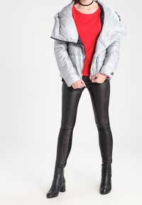 Veste matelassée argentée avec un col oversize, portée sur un haut rouge, associée à des leggings en cuir noir et des bottines. Textures lisses et design élégant.