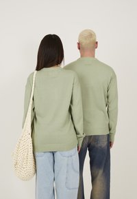 Grønne strikkede trøjer med lange ærmer, rund hals og ribbet nederkant, sammen med løse, lyseblå og mørkeblå jeans samt en beige hæklede taske.