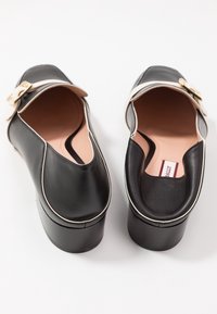 Mocassins à plateforme en cuir noir avec liseré blanc, brides à boucle dorée et semelles intérieures roses sur fond blanc.