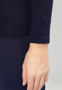 Zbliżenie na rękę spoczywającą obok, ubraną w ciemnoniebieską bluzę z długim rękawem i prążkowanymi mankietami.