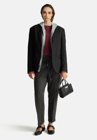 Žena modelující vrstvený outfit s černým blejzrem, šedou mikinou na zip, vínovým topem, tmavě šedými kalhotami, černými balerínami a držící malou černou kabelku.