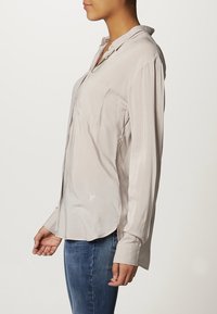 Vaaleanbeige pitkähihaisen paidan sileä kangas, jossa on kaulus, kaksi etutaskua ja kaareva helma. Sivulla hienovarainen logokoriste.