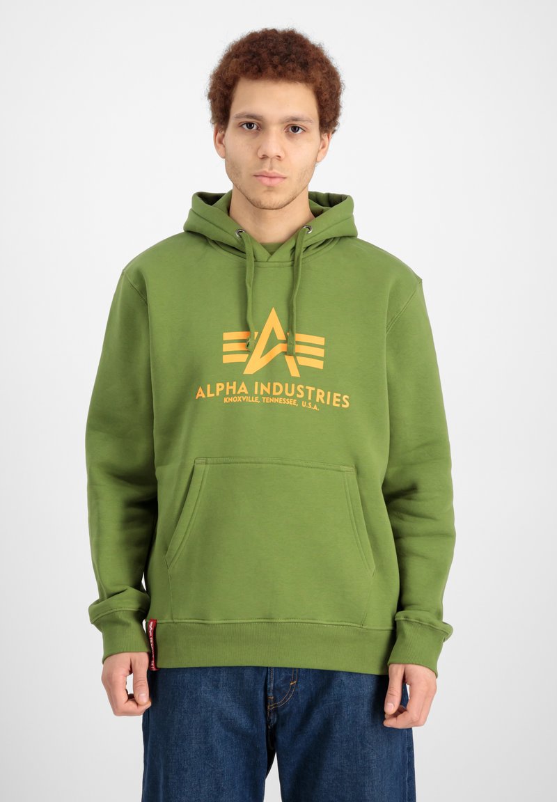 Zöld kapucnis pulóver elülső zsebbel és sárga nyomott logóval, amelyen az "ALPHA INDUSTRIES" felirat olvasható. Puha, texturált anyagból készült, laza szabású.