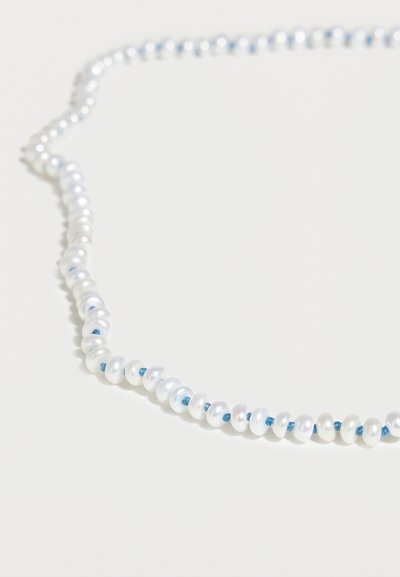Μονόκλωνο κολιέ με μικρές λευκές πέρλες περασμένες σε μπλε κορδόνι, τοποθετημένο σε καμπυλωτή γραμμή πάνω σε λευκή επιφάνεια.