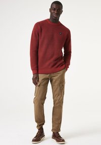 Roter strukturierten Strickpullover mit Rundhalsausschnitt, kombiniert mit beigen Cargo-Hosen mit Seitentaschen und braunen Schnürstiefeln.