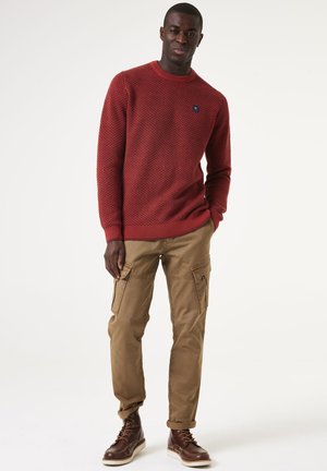 Roter strukturierten Strickpullover mit Rundhalsausschnitt, kombiniert mit beigen Cargo-Hosen mit Seitentaschen und braunen Schnürstiefeln.