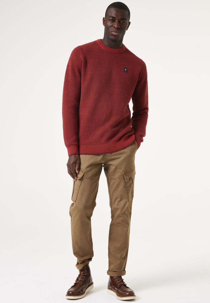 Roter strukturierten Strickpullover mit Rundhalsausschnitt, kombiniert mit beigen Cargo-Hosen mit Seitentaschen und braunen Schnürstiefeln.