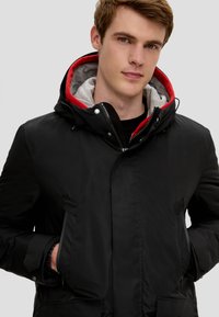 Schwarze gepolsterte Jacke mit Reißverschluss und Druckknopfverschluss, ausgestattet mit einem roten und grauen Kapuzenfutter, zwei Fronttaschen und elastischen Bündchen.