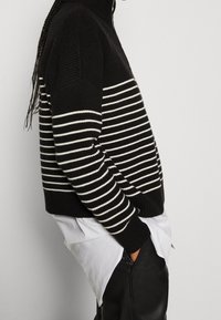 Schwarz-weiß gestreifter Pullover mit hohem Kragen, gerippter Textur und überschnittenen Schultern, kombiniert mit einem weißen Hemd und schwarzen Hosen.