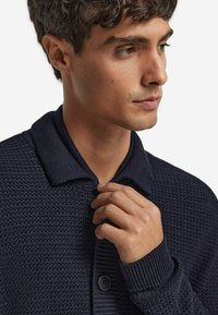 Marinblå stickad cardigan med ett strukturerat mönster, med krage, knappdetalj och ribbade manschetter. Närbild av halsringningen och handen.