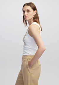 Top de tirantes blanco con un patrón decorativo de tachuelas de color dorado; pantalones beige con detalles de tachuelas a lo largo de los bolsillos.