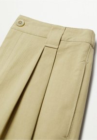 Un pantalon plissé beige présente un tissu lisse avec une fermeture à bouton sur le devant, des poches latérales et une taille soigneusement cousue.
