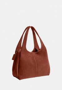 Sac à main en daim marron avec double poignée, bords supérieurs arrondis et petite étiquette attachée sur un fond neutre.
