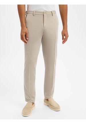 Mann trägt beige Slim-Fit-Hosen und passende Slipper, steht vor einem schlichten weißen Hintergrund.