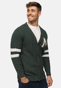 Groene kabelgebreide cardigan met witte strepen op de mouwen en een geborduurde letter "M." Heeft een V-hals en knoopsluiting. Zachte textuur.