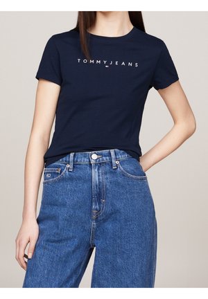 Kobieta w granatowej koszulce Tommy Jeans i wysokich niebieskich dżinsach, stojąca z jedną ręką na biodrze.