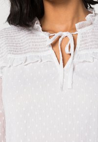 White blouse with a textured dot pattern, ruffled neckline, and a tie closure. Soft, lightweight fabric with a crinkled surface.