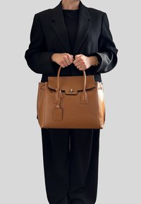 Brązowa skórzana torebka o strukturalnym designie, z przednim klapkiem, górnymi uchwytami i detalem w postaci metki bagażowej. Gładka tekstura i minimalistyczne okucia.