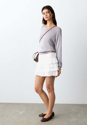 Mlada žena u svijetlopurpurnoj majici dugih rukava, bijeloj naboranoj mini suknji, smeđoj torbi preko tijela i tamnosmeđim balerinkama, stoji na sivoj podlozi.