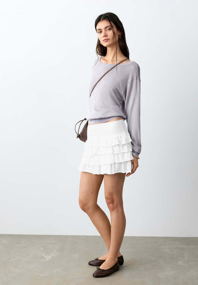 Jeune femme portant un haut à manches longues violet clair, une mini-jupe blanche à volants, un sac bandoulière marron et des ballerines marron foncé, debout sur un sol gris.