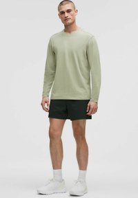 Hellgrünes Langarmshirt mit rundem Ausschnitt, kombiniert mit schwarzen Shorts. Das Modell trägt weiße Sportschuhe und Socken und steht aufrecht.