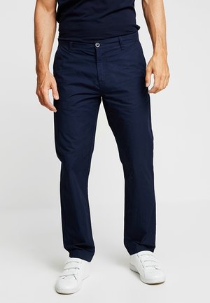 Mann trägt dunkelblaue gerade geschnittene Hose und weiße Klettverschluss-Sneaker, steht vor einem schlichten hellen Hintergrund.