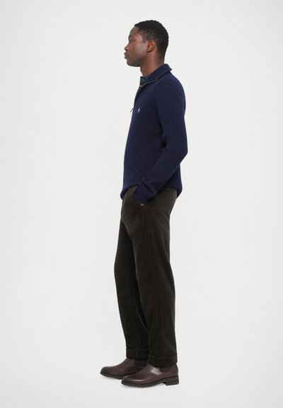 Pulover navy cu pliuri, cu guler, asortat cu pantaloni din corduroy închis și cizme din piele vișinii. Croiala este relaxată, cu un detaliu subtil al logo-ului.