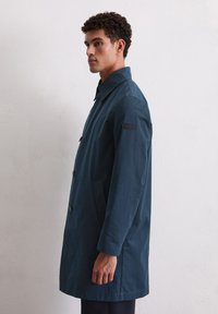 Mann mit lockigem Haar, der einen dunkelblauen Knopfmantel trägt und im Profil vor einer schlichten weißen Wand steht.