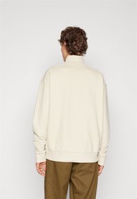 Sudadera beige con cuello alto, ajuste holgado, hombros caídos y puños acanalados, combinada con pantalones verde oliva.