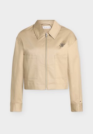Veste courte beige avec fermeture éclair intégrale à l'avant, col pointu, deux poches avant et petit logo brodé sur la poitrine gauche et le poignet.