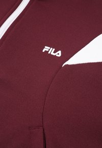 Maroon huppari valkoisilla yksityiskohdilla, jossa on vetoketjusuljenta ja kohotettu FILA-logo valkoisena edessä. Pehmeä, teksturoitu kangas.