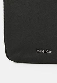 Borsa nera in tessuto con superficie texturizzata, caratterizzata dal logo "Calvin Klein" in argento. Design slim con bordi arrotondati e dettagli cuciti.