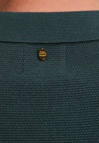 Suéter de punto texturizado en verde oscuro con cuello acanalado, que presenta una etiqueta de logo PME Legend en negro y dorado sujeta por costuras naranjas en la parte posterior.