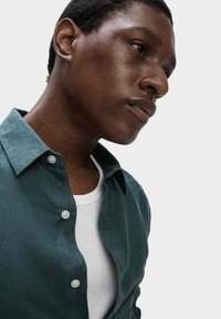 Camisa de botones en lino verde oscuro con cuello y dos botones blancos. Lleva una camiseta blanca de cuello redondo por debajo. Textura suave.
