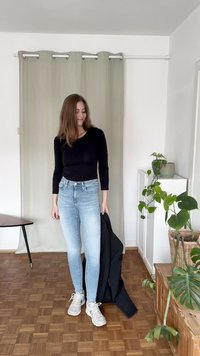 Top de manga longa preto, jeans skinny azul claro, ténis cinza, segurando um blazer preto riscado. Ambiente interno com plantas e chão de madeira clara.