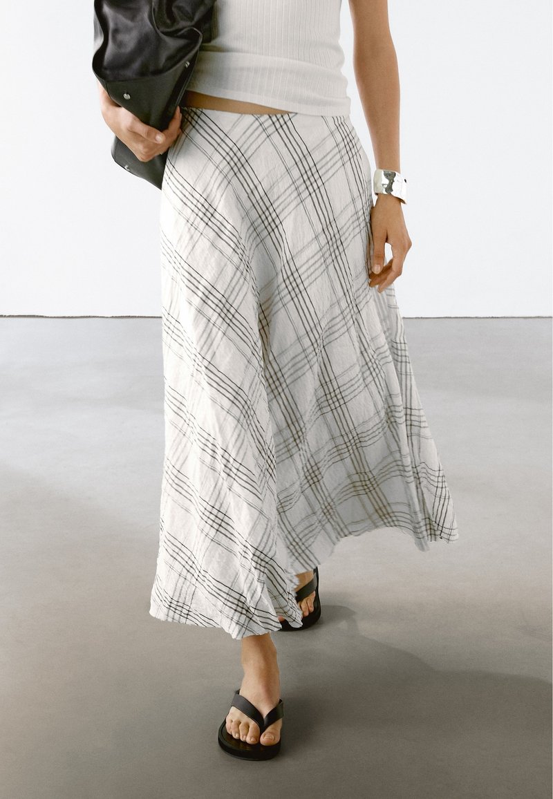 Femme portant un haut blanc sans manches et une longue jupe blanche avec des lignes de grille noires, des sandales noires, un bracelet manchette argenté, tenant un sac noir.