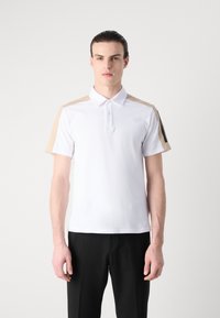 Balta polo marškiniai su smėlio spalvos peties akcentais, trumpomis rankovėmis ir klasikiniais klostais. Audinys atrodo lygus ir kvėpuojantis.