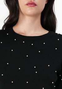Pull noir avec un col rond, en tissu texturé, décoré de perles blanches éparpillées sur toute la surface.