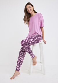 Jasnofioletowa bluzka z krótkim rękawem w połączeniu z dopasowanymi fioletowymi spodniami w kwiatowy wzór. Modelka siedzi na białym stołku, boso.