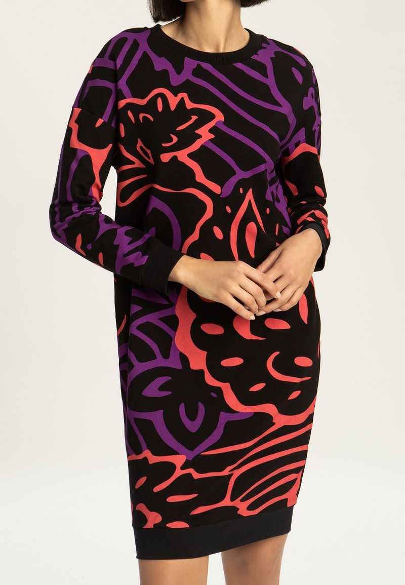 Femme portant une robe noire à manches longues avec des motifs floraux abstraits rouges et violets audacieux, les mains doucement jointes à la taille.