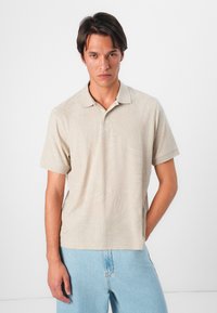 Beige πόλο μπλούζα με κοντά μανίκια και κολάρο, σε συνδυασμό με έναν διακριτικό σχέδιο φύλλου. Φορεμένη με ανοιχτόχρωμα τζιν παντελόνια.