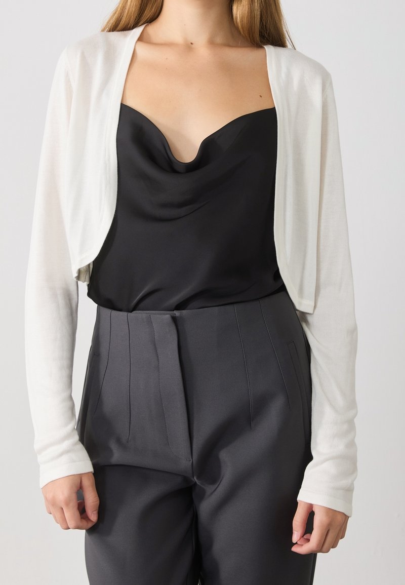 Femme portant un cardigan court blanc par-dessus un caraco noir drapé, rentré dans un pantalon gris foncé cintré taille haute.