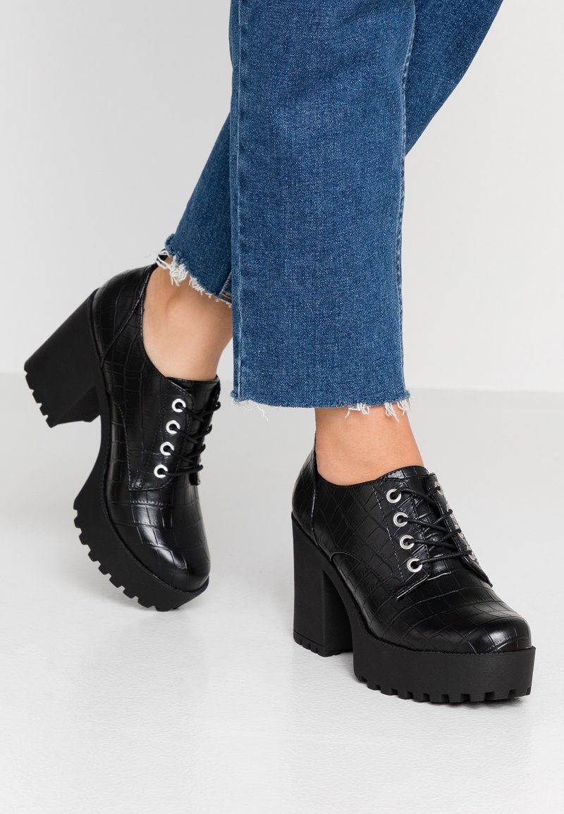 Jambes portant un jean bleu à ourlet effiloché et des chaussures à plateforme noires épaisses à lacets avec un motif texturé sur un sol blanc.