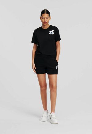 Black T-shirt with a pocket and white graphic detail, paired with black shorts. Model wearing white sneakers. Neutral gray background.