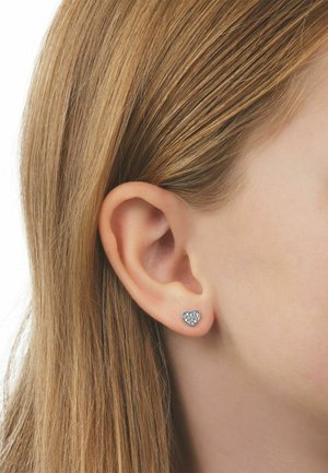 Orecchio di donna con capelli biondi lisci raccolti dietro, con un piccolo orecchino d'argento a forma di cuore con pietre chiare incastonate.